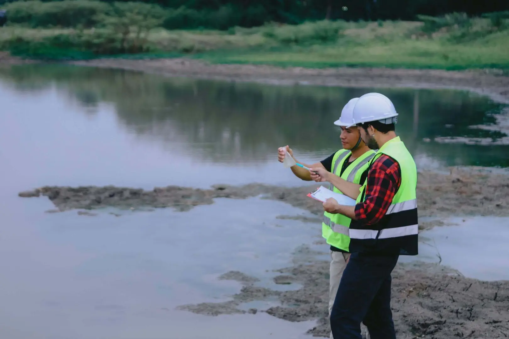 Two workers in safety vests and helmets inspecting a water body.