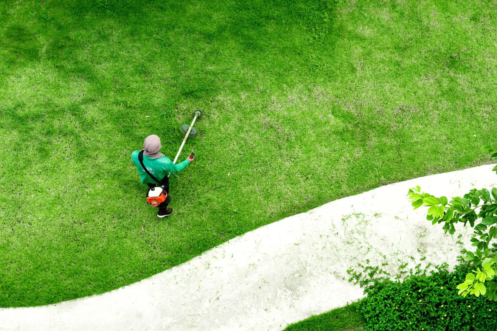 Aerial view of a person in a green shirt using a grass trimmer on a bright green lawn, near a curving white path with leafy greenery nearby.