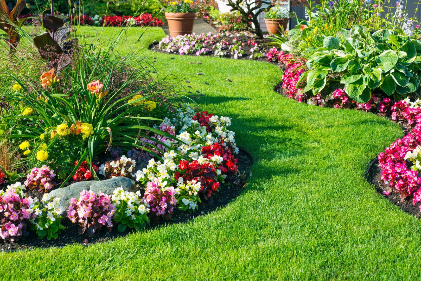A vibrant garden with lush green grass and curved flower beds bursting with colorful flowers, including pink, red, and yellow blooms, creating a serene atmosphere.